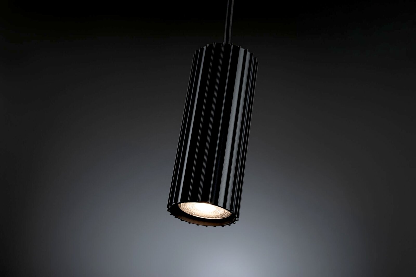 Schwarze, zylindrische LED-Pendelleuchte mit geriffeltem Lampenschirm und warmweiß leuchtendem Spot, schräg von unten im 3/4-Winkel vor dunklem Hintergrund
