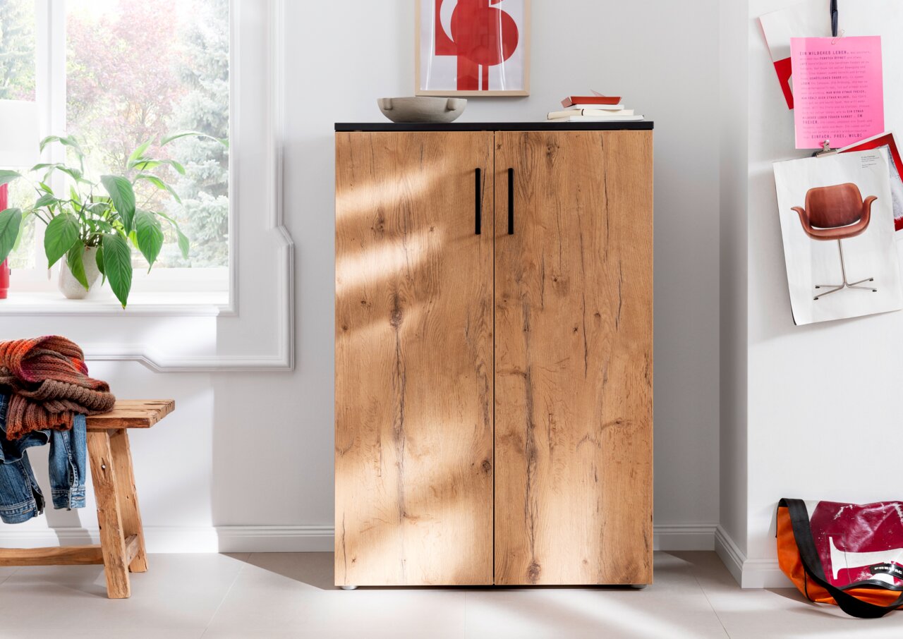 Schuhschrank aus Holz mit zwei Türen, schwarze Griffe, in einem hellen Raum mit Pflanzen und Dekorationen, Frontalansicht