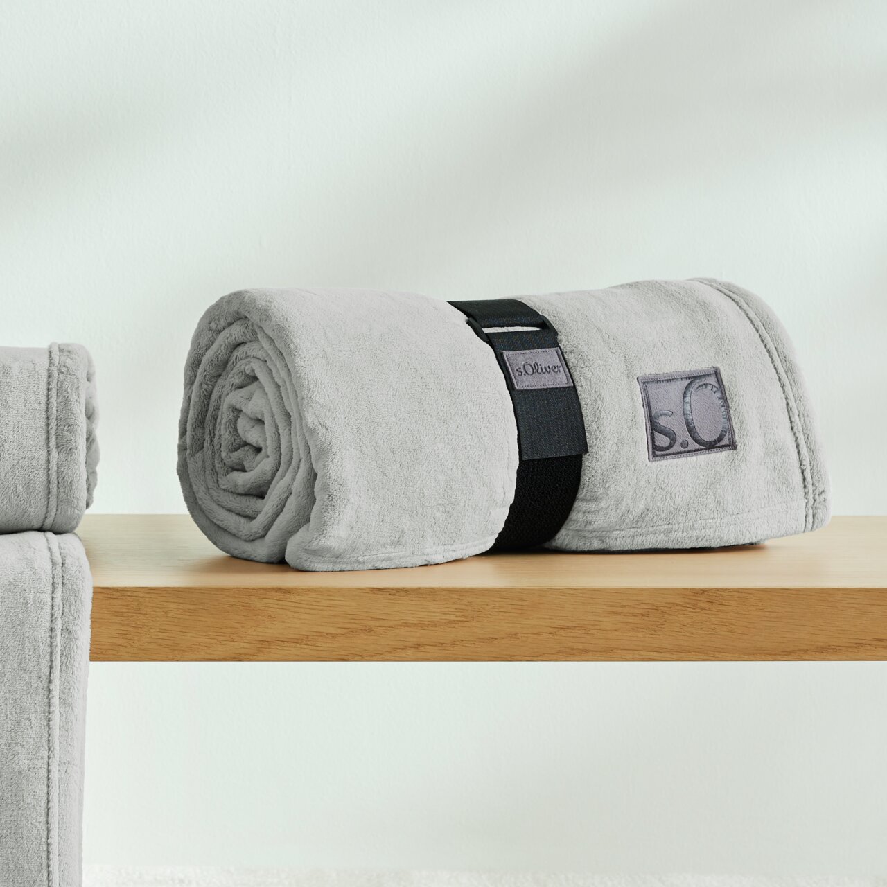 Graue, gerollte Kuscheldecke von s.Oliver auf einem Holztisch, seitliche Perspektive.