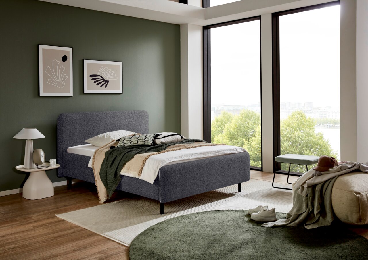 Modernes Polsterbett in einem stilvollen Schlafzimmer mit großen Fenstern, seitliche Perspektive.