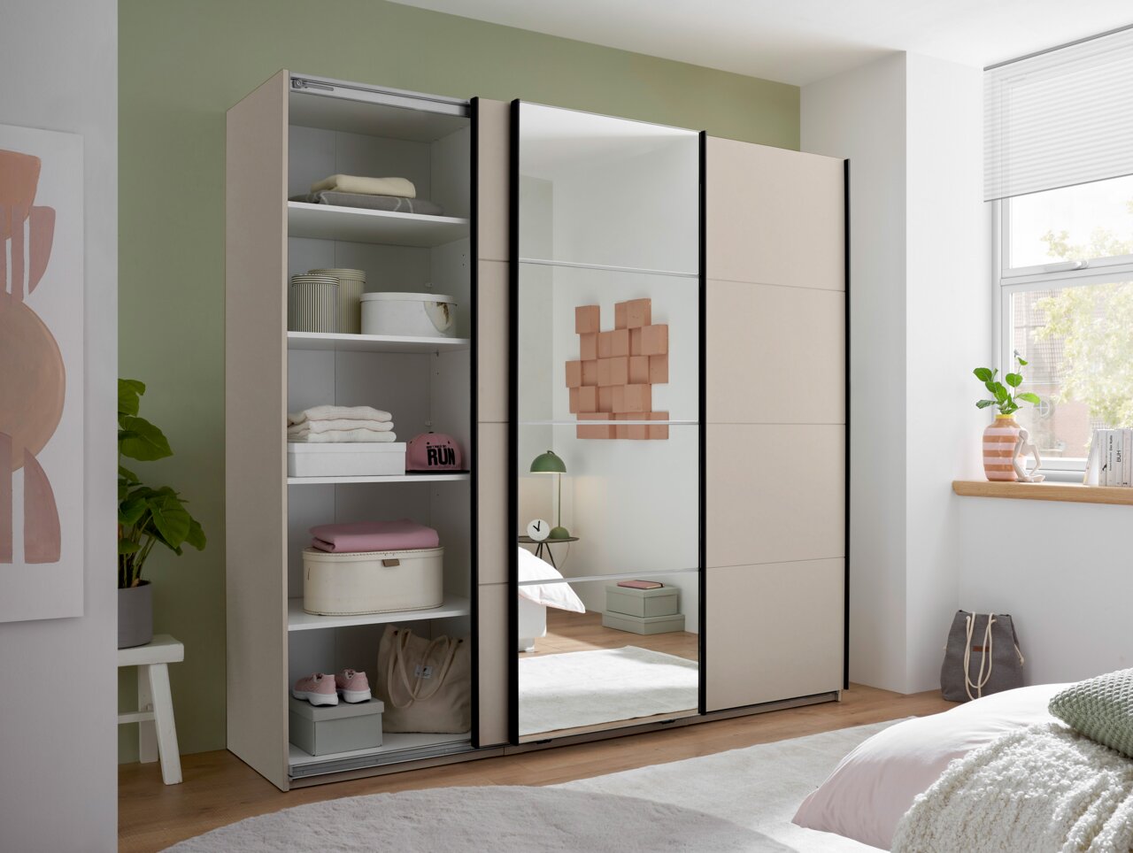 Beiger Schwebetürenschrank im Schlafzimmer, Frontansicht leicht schräg von rechts; linke Schiebetür geöffnet mit sichtbaren Regalböden, mittlere Tür mit großem Spiegel, rechte Tür geschlossen.