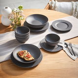 Ein stilvolles, 16-teiliges Geschirrset in mattem Schwarz auf einem Holztisch, bestehend aus Tellern, Schalen und Tassen, mit einer modernen, strukturierten Oberfläche. Perspektive von oben.