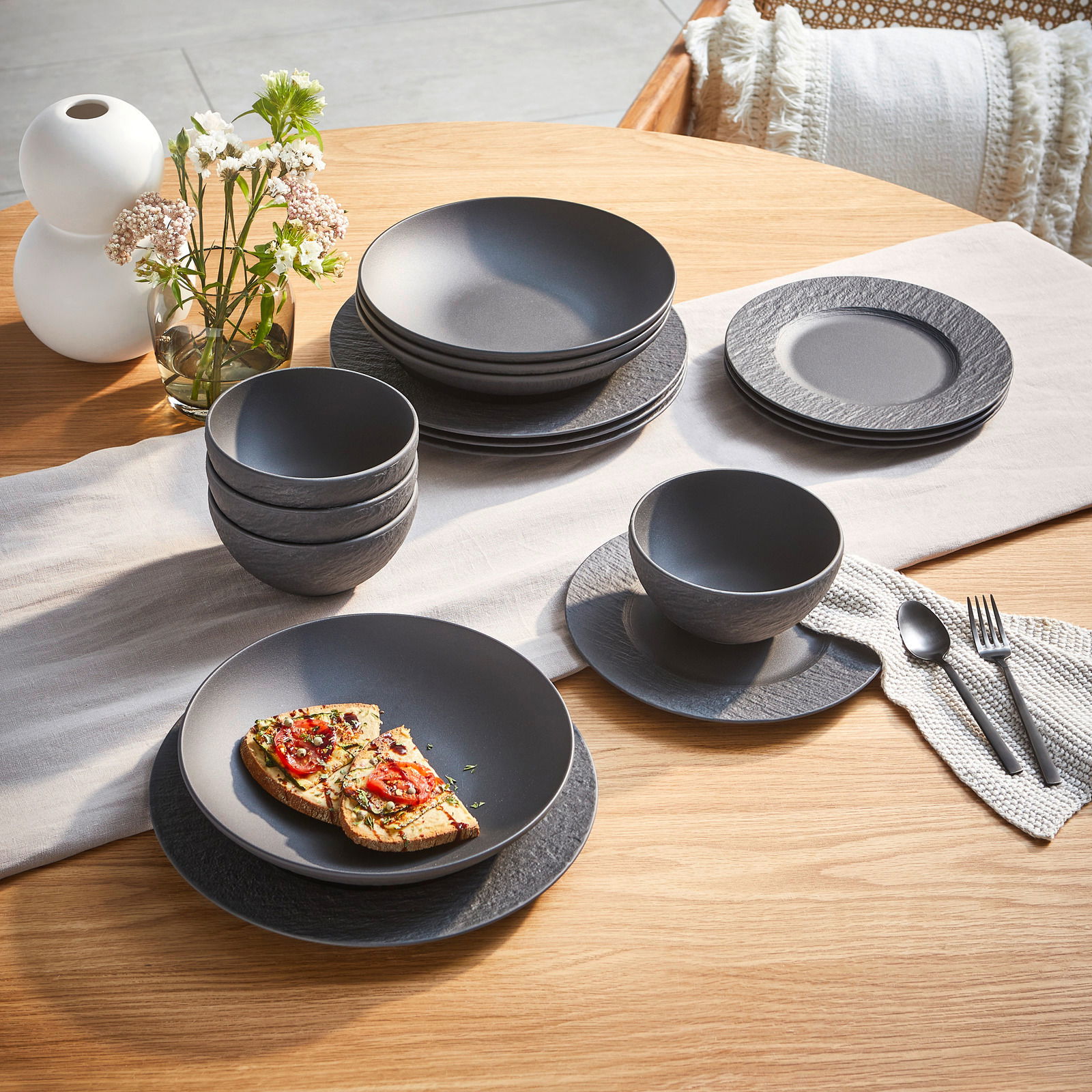 Ein stilvolles, 16-teiliges Geschirrset in mattem Schwarz auf einem Holztisch, bestehend aus Tellern, Schalen und Tassen, mit einer modernen, strukturierten Oberfläche. Perspektive von oben.