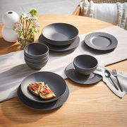 Ein stilvolles, 16-teiliges Geschirrset in mattem Schwarz auf einem Holztisch, bestehend aus Tellern, Schalen und Tassen, mit einer modernen, strukturierten Oberfläche. Perspektive von oben.