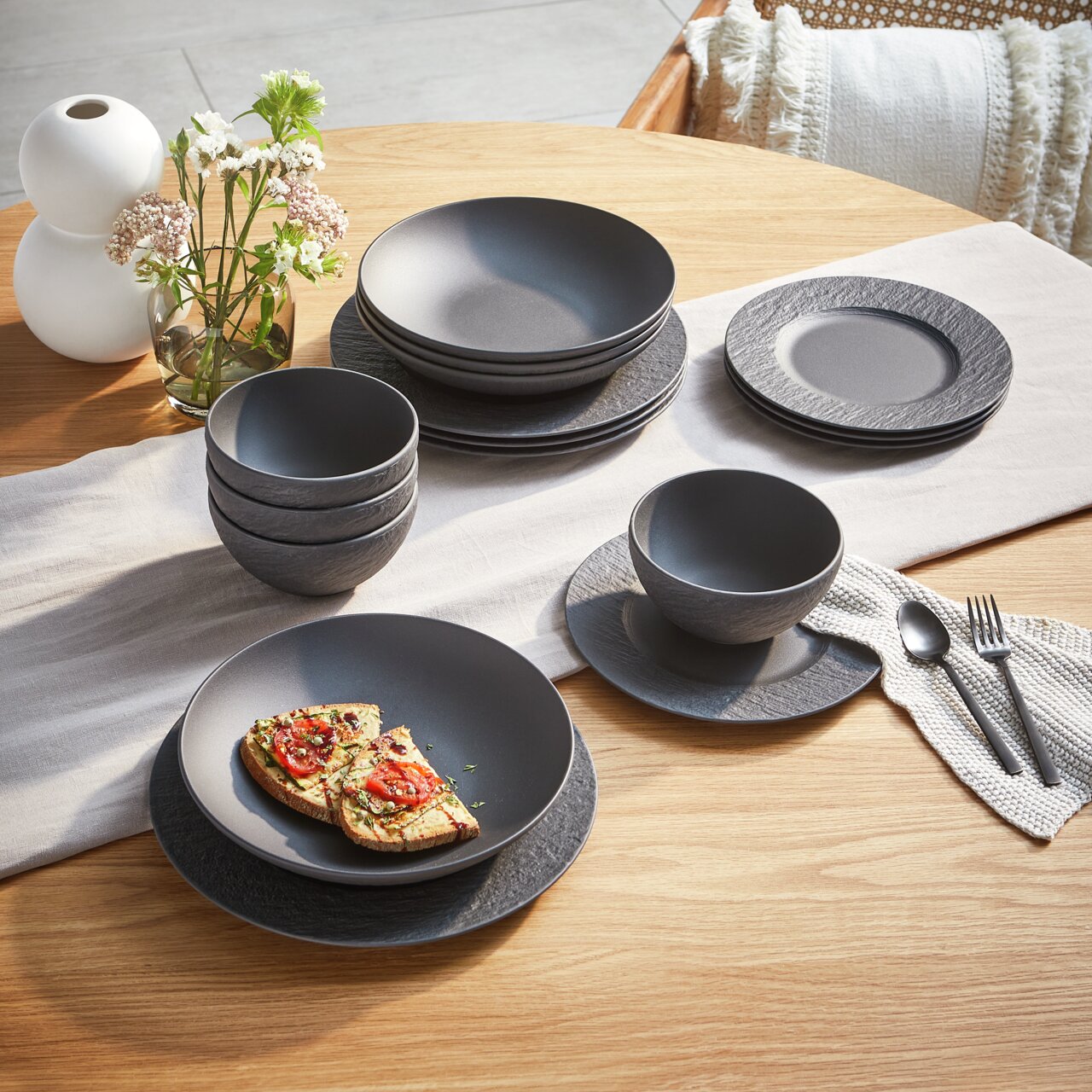 Een stijlvolle, 16-delige servieset in matzwart op een houten tafel, bestaande uit borden, schalen en kopjes, met een modern, gestructureerd oppervlak. Aanzicht van bovenaf.