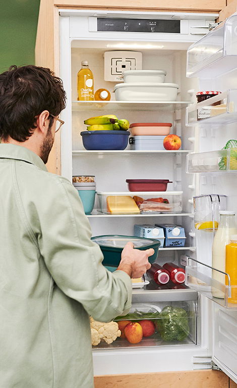 Ein geöffneter Kühlschrank mit mehreren bunten Schüsseln, darunter die Multischüssel Vivid Mauve, die von einem Mann in einem grünen Hemd betrachtet wird. Perspektive von hinten auf den Mann und den Kühlschrank.