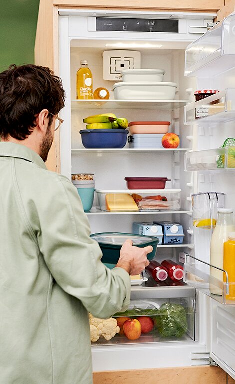 Ein geöffneter Kühlschrank mit mehreren bunten Schüsseln, darunter die Multischüssel Vivid Mauve, die von einem Mann in einem grünen Hemd betrachtet wird. Perspektive von hinten auf den Mann und den Kühlschrank.