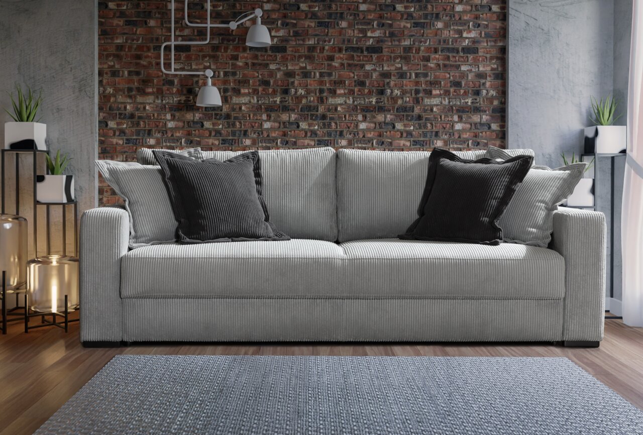 Graues Querschläfer-Sofa mit schwarzen Kissen, frontal vor einer Ziegelwand fotografiert.