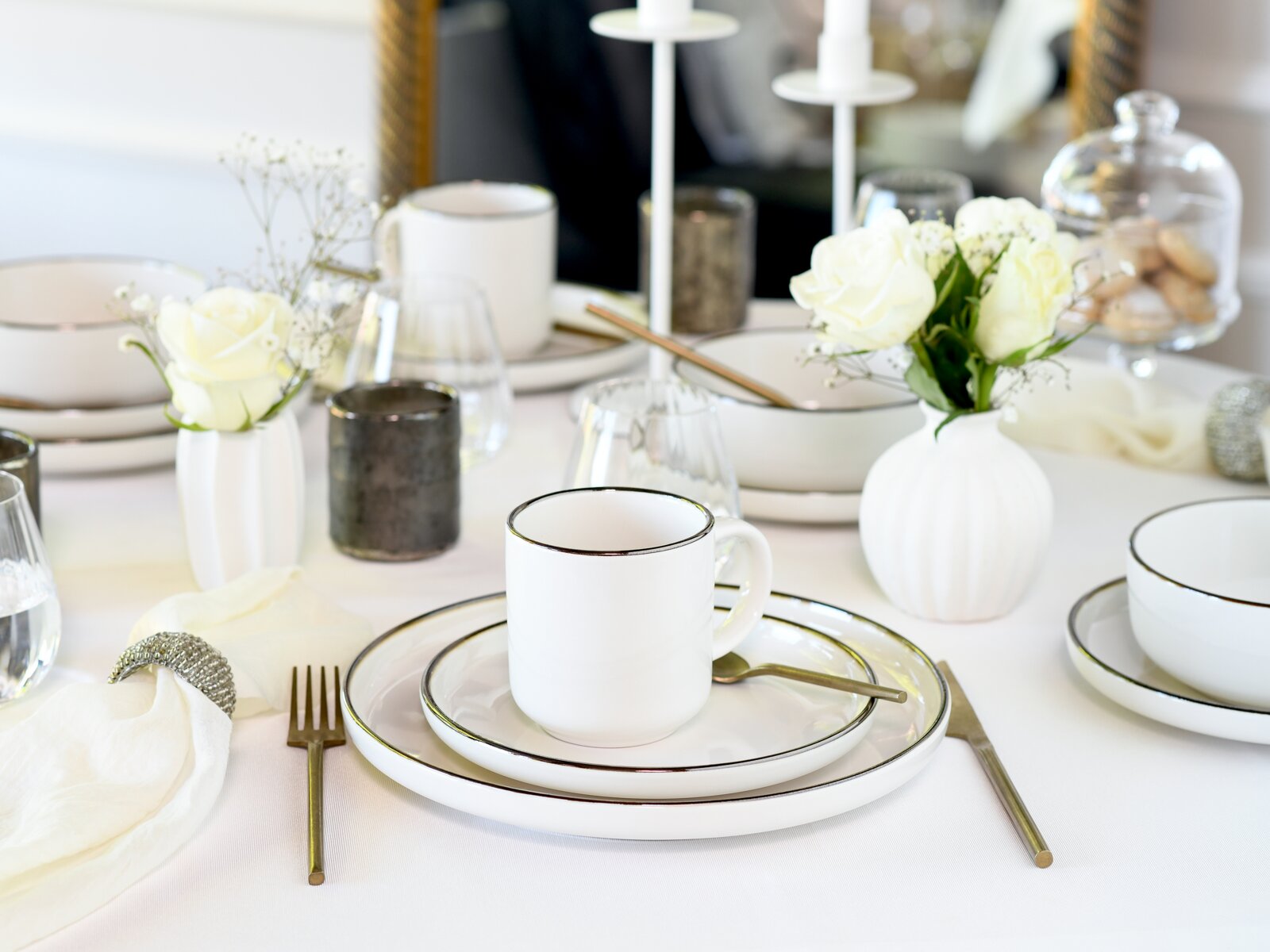 Elegant gedeckter Tisch mit weißem Geschirr mit feinem dunklem Rand, Tasse auf Tellern, goldfarbenem Besteck, Gläsern und weißen Rosen in Vasen, fotografiert aus leicht erhöhter schräger Perspektive.