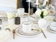 Elegant gedeckter Tisch mit weißem Geschirr mit feinem dunklem Rand, Tasse auf Tellern, goldfarbenem Besteck, Gläsern und weißen Rosen in Vasen, fotografiert aus leicht erhöhter schräger Perspektive.