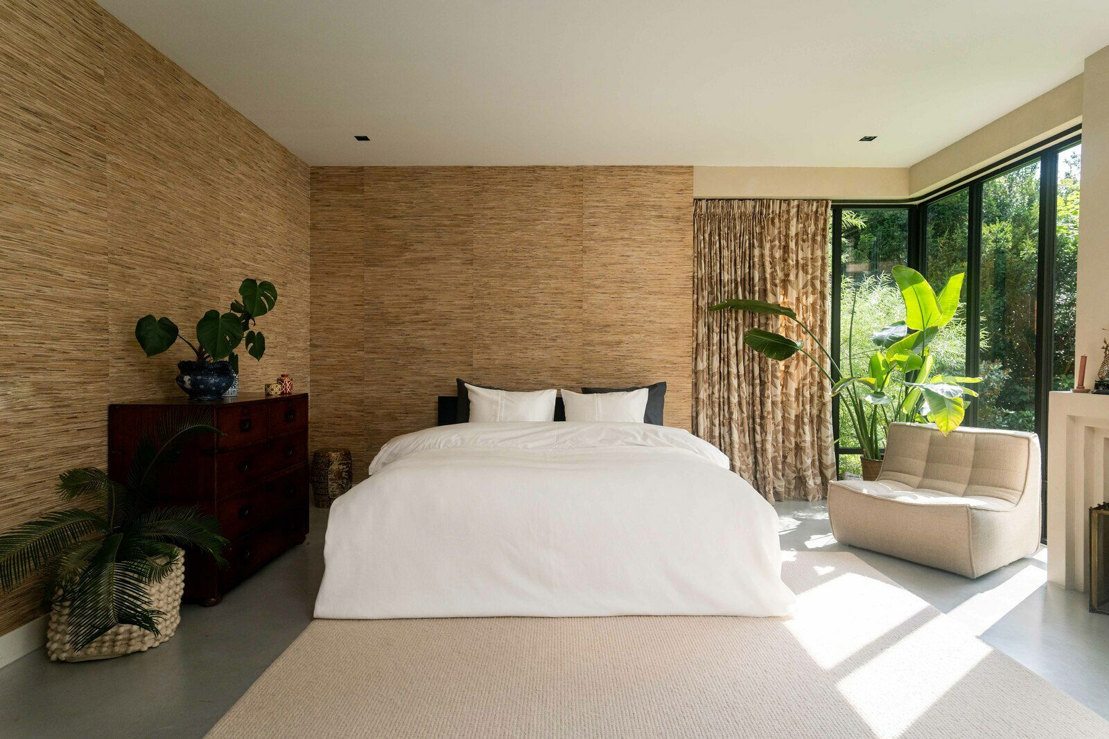 Modernes Schlafzimmer mit weißer Bettwäsche auf einem Doppelbett, umgeben von Pflanzen und natürlichem Licht, aus der Frontperspektive.