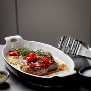 Weiße ovale Auflaufform mit Griffen, gefüllt mit Steak in Sauce und Tomaten sowie Kräutern, in schräger Seitenansicht von vorne auf dunkler Arbeitsfläche