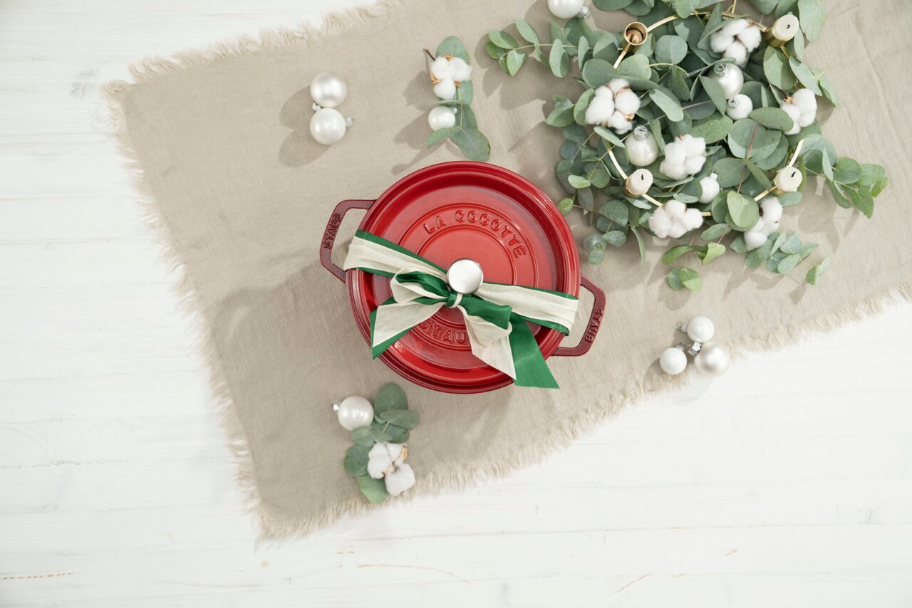 Rote runde Cocotte mit Schleife, von oben fotografiert, auf einem beigen Tuch mit Dekoration aus Eukalyptus und Baumwolle.