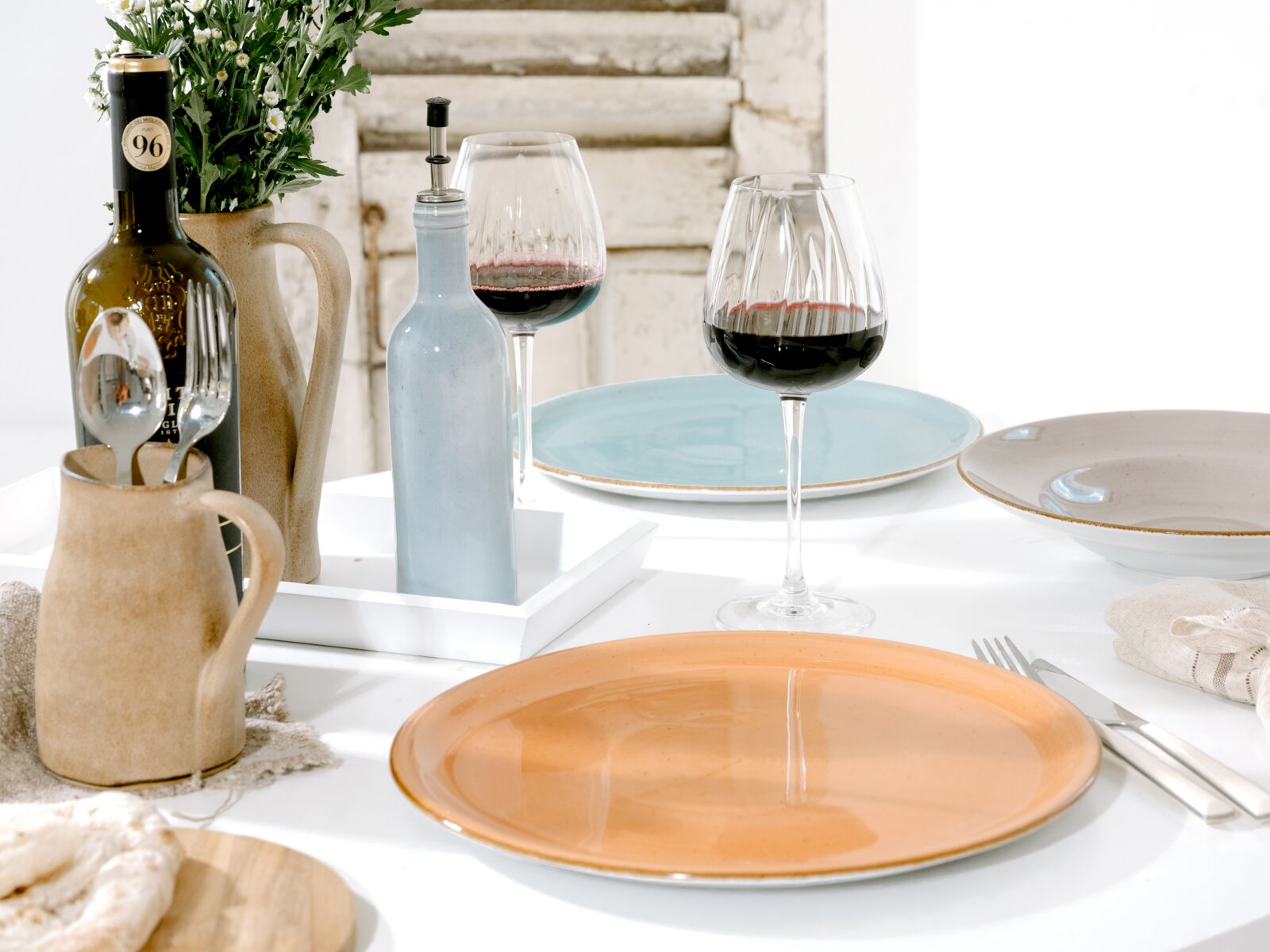 Gedeckter Esstisch mit farbigen Keramiktellern (orange, blau, beige), zwei Rotweingläsern, Weinflasche, hellblauer Flasche und Krug mit Blumen; Perspektive: leicht erhöht aus schräger Vorderansicht.