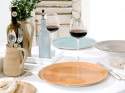Gedeckter Esstisch mit farbigen Keramiktellern (orange, blau, beige), zwei Rotweingläsern, Weinflasche, hellblauer Flasche und Krug mit Blumen; Perspektive: leicht erhöht aus schräger Vorderansicht.