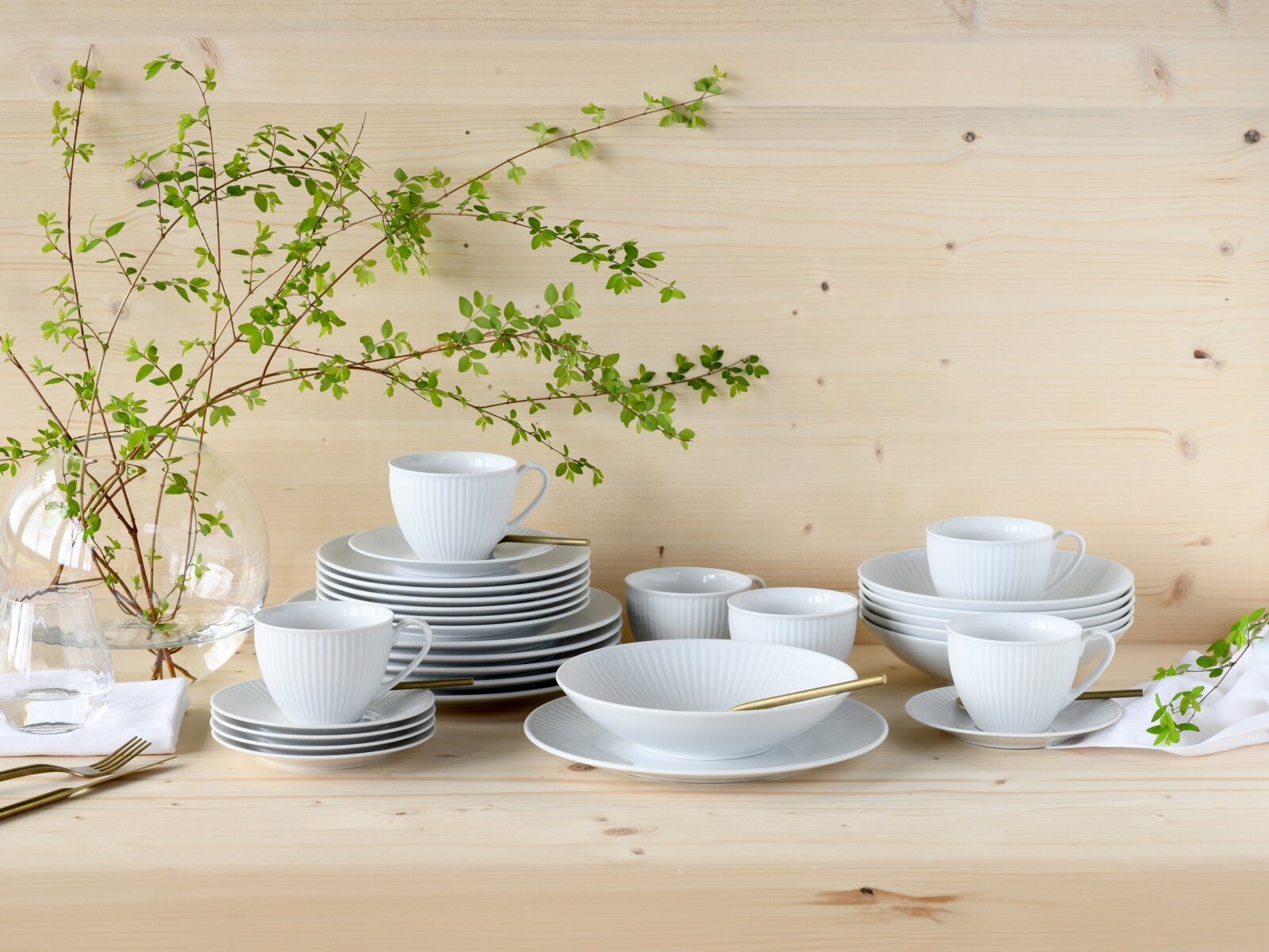 Creatable Kombiservice 30-tlg VALENCIA Weißes geriffeltes Porzellan-Geschirrset mit Tassen, Tellern und Schalen auf hellem Holztisch, dekoriert mit grünen Zweigen und Glasvase, in frontaler Tisch-Perspektive