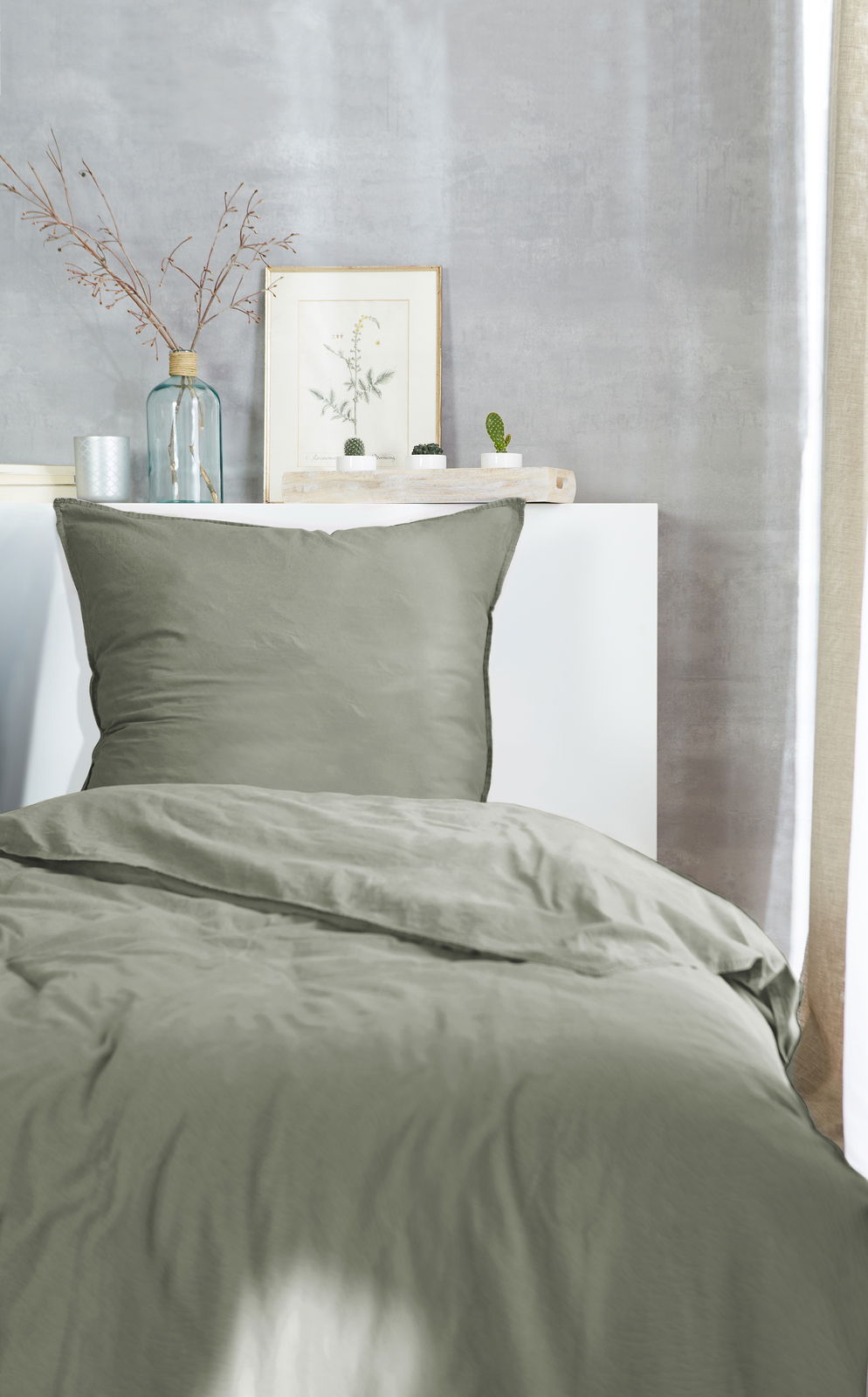 Bettwäsche in Olivgrün auf einem Bett, fotografiert aus der Perspektive vom Fußende. Im Hintergrund sind eine Vase mit Zweigen, ein gerahmtes Bild und kleine Pflanzen zu sehen.