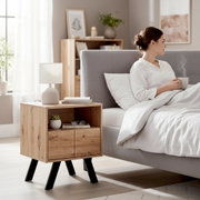 Moderner Nachttisch aus Holz mit offenem Fach und zwei Türen auf schwarzen Beinen neben grauem Polsterbett, seitliche Perspektive im hellen Schlafzimmer