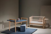 Ein modernes Kinderzimmer-Set bestehend aus einem blauen Tisch mit Holzbeinen und einem passenden Hocker, sowie einem hellen Kinderbett mit Gitterstäben. Die Perspektive zeigt das Set von vorne in einem minimalistisch eingerichteten Raum.