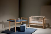 Ein modernes Kinderzimmer-Set bestehend aus einem blauen Tisch mit Holzbeinen und einem passenden Hocker, sowie einem hellen Kinderbett mit Gitterstäben. Die Perspektive zeigt das Set von vorne in einem minimalistisch eingerichteten Raum.