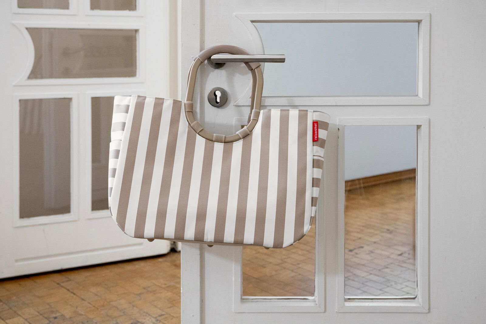 Gestreifte Tragetasche in Beige und Weiß mit runden Henkeln, an einer weißen Tür am Türgriff hängend, Frontansicht auf Augenhöhe