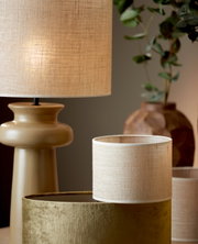 Cilindrische lampenkap in naturel beige, op de voorgrond op een tafel geplaatst, met een stijlvolle lamp en planten op de achtergrond.