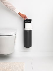 Moderner Toilettenrollenspender in schwarzer Farbe, an der Wand montiert, mit einer Hand, die den Deckel öffnet. Seitenansicht.