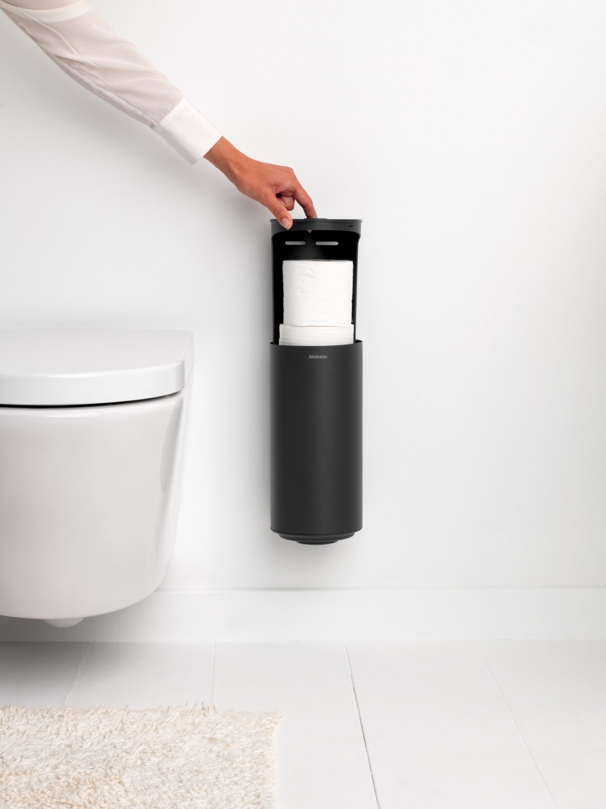 Moderner Toilettenrollenspender in schwarzer Farbe, an der Wand montiert, mit einer Hand, die den Deckel öffnet. Seitenansicht.