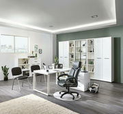 Moderne Büroumgebung mit weißem Eck-Schreibtisch, schwarzem Bürostuhl und Regalen im Hintergrund, seitliche Perspektive.