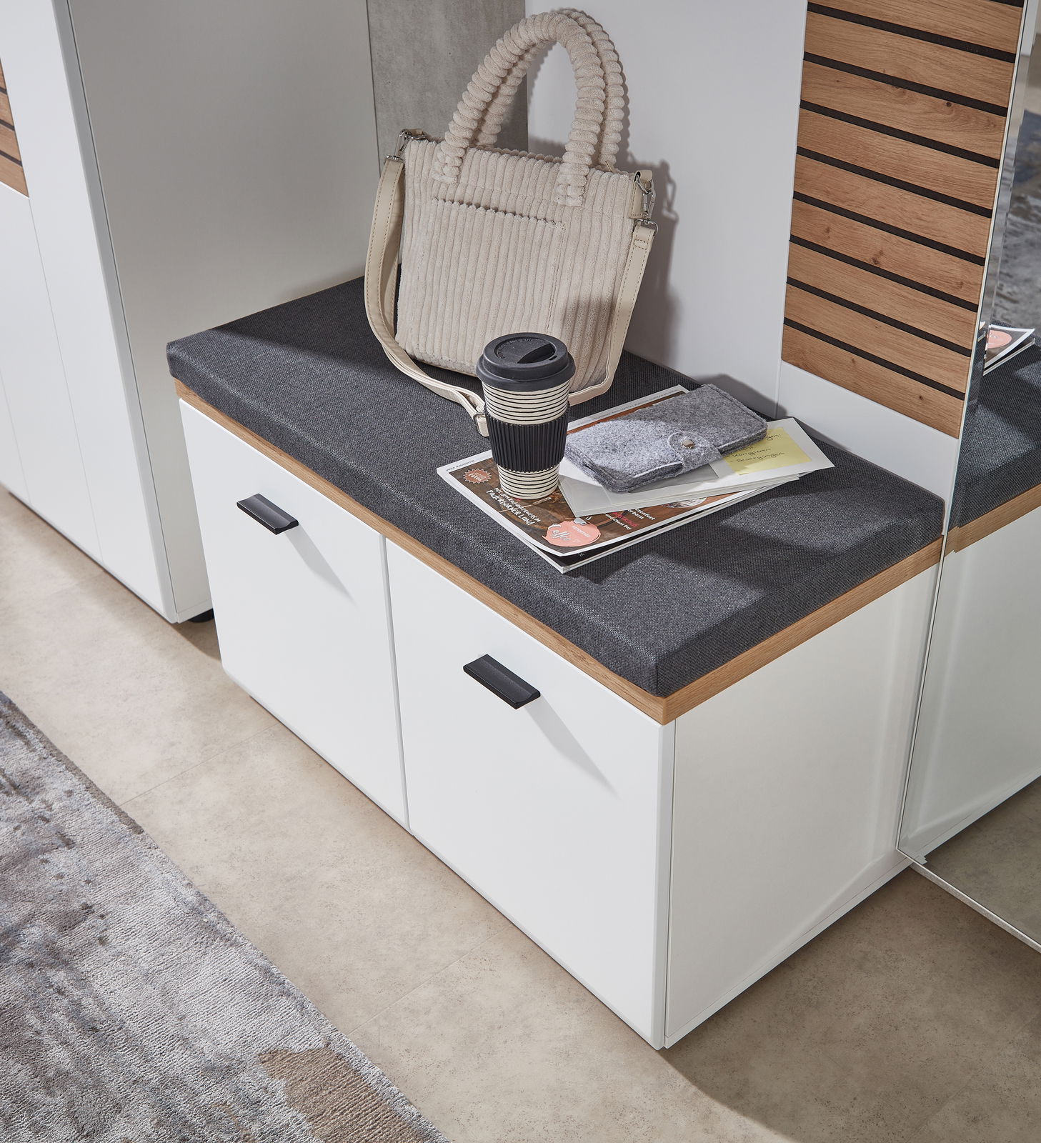 Garderobenbank mit weißem Korpus und grauem Sitzpolster, darauf eine beige Tasche und ein Kaffeebecher, aus der Vogelperspektive fotografiert.