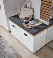 Garderobenbank mit weißem Korpus und grauem Sitzpolster, darauf eine beige Tasche und ein Kaffeebecher, aus der Vogelperspektive fotografiert.