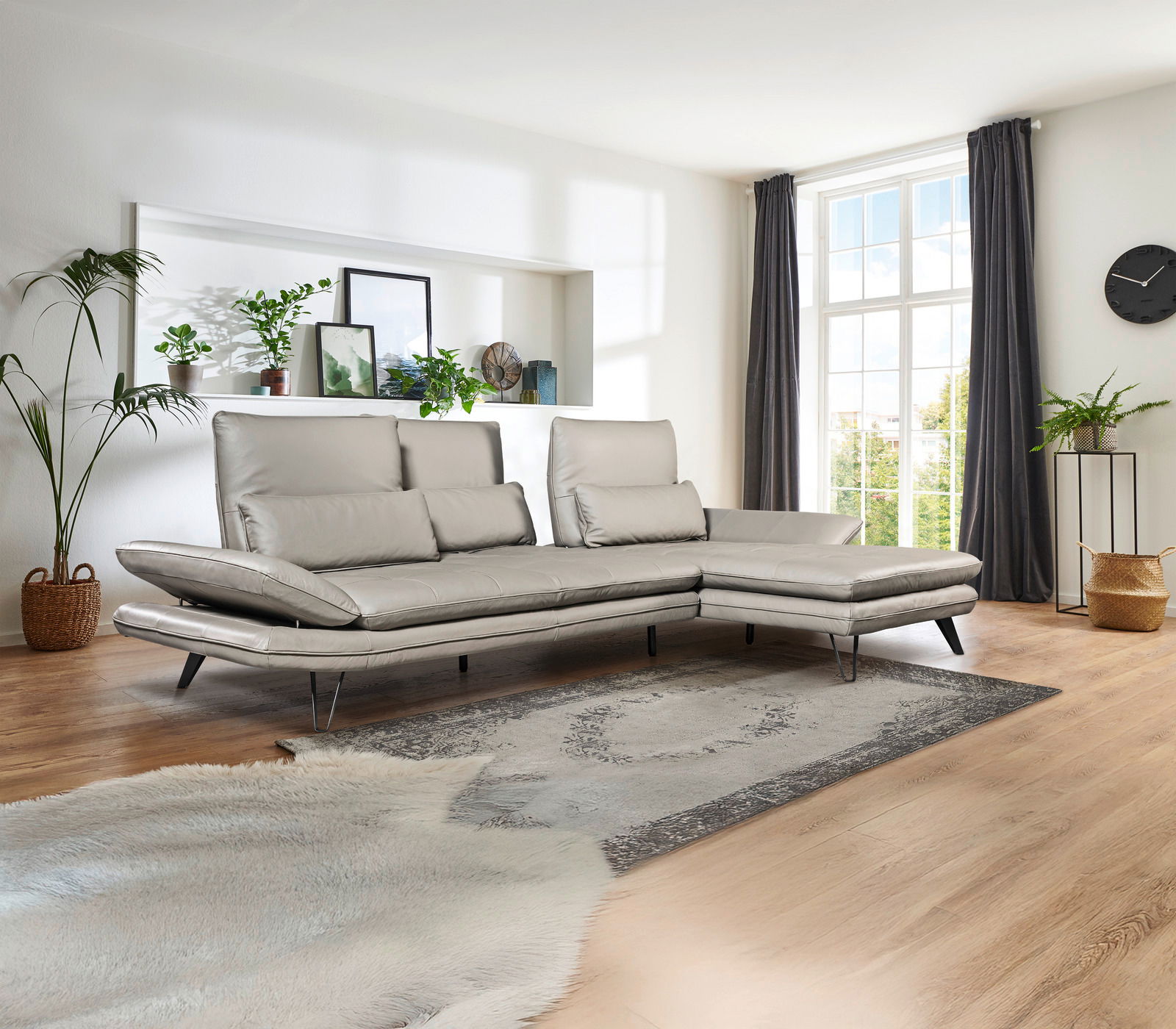 Moderne Longchairkombination in einem hellen Wohnzimmer mit großen Fenstern, grauen Vorhängen und Pflanzen, seitliche Perspektive.