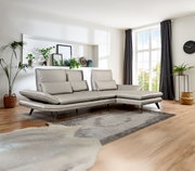 Moderne Longchairkombination in einem hellen Wohnzimmer mit großen Fenstern, grauen Vorhängen und Pflanzen, seitliche Perspektive.