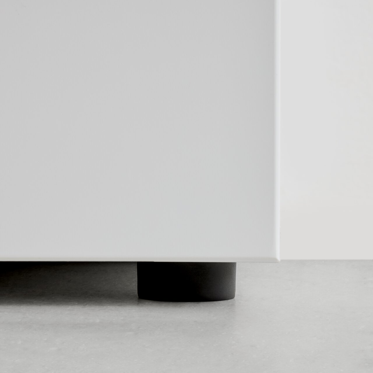 Nahaufnahme der unteren Ecke eines weißen Garderobenschranks mit schwarzem Fuß auf grauem Boden.