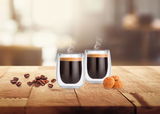 Zwei doppelwandige Thermogläser mit heißem Espresso, umgeben von Kaffeebohnen und Keksen, auf einem Holztisch in einem gemütlichen Ambiente, seitliche Perspektive.