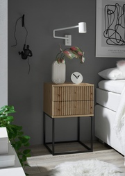 Moderne Nachtkommode aus Holz mit gerillter Front und schwarzem Metallgestell, seitliche Perspektive, dekoriert mit einer Vase und einer Uhr.
