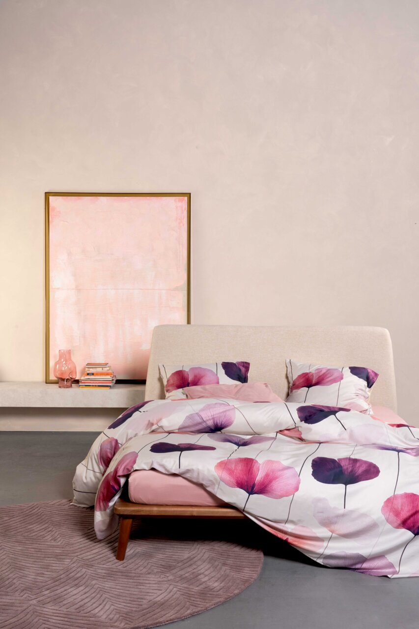 Modernes Schlafzimmer mit Polsterbett und Bettwäsche mit großen rosa‑ und lilafarbenen Blüten auf weißem Grund, frontal aus leicht erhöhter Perspektive fotografiert im Hintergrund ein gerahmtes Bild, Bücher und eine rosa Vase.