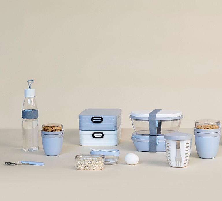 Verschiedene hellblaue und weiße Frischhalteboxen und Behälter, darunter eine Wasserflasche, stapelbare Brotdosen und runde Behälter, auf einer neutralen Oberfläche aus der Frontalperspektive.