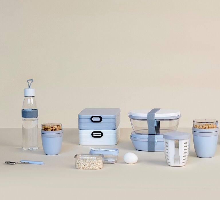 Verschiedene hellblaue und weiße Frischhalteboxen und Behälter, darunter eine Wasserflasche, stapelbare Brotdosen und runde Behälter, auf einer neutralen Oberfläche aus der Frontalperspektive.