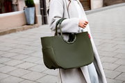 Olivgrüne Tote-Bag/Handtasche aus strukturiertem Stoff mit runden Griffen, seitlicher Außentasche und kleinem roten Label, getragen am Unterarm einer Person im hellgrauen Mantel; Perspektive: seitliche Nahaufnahme auf die Tasche.
