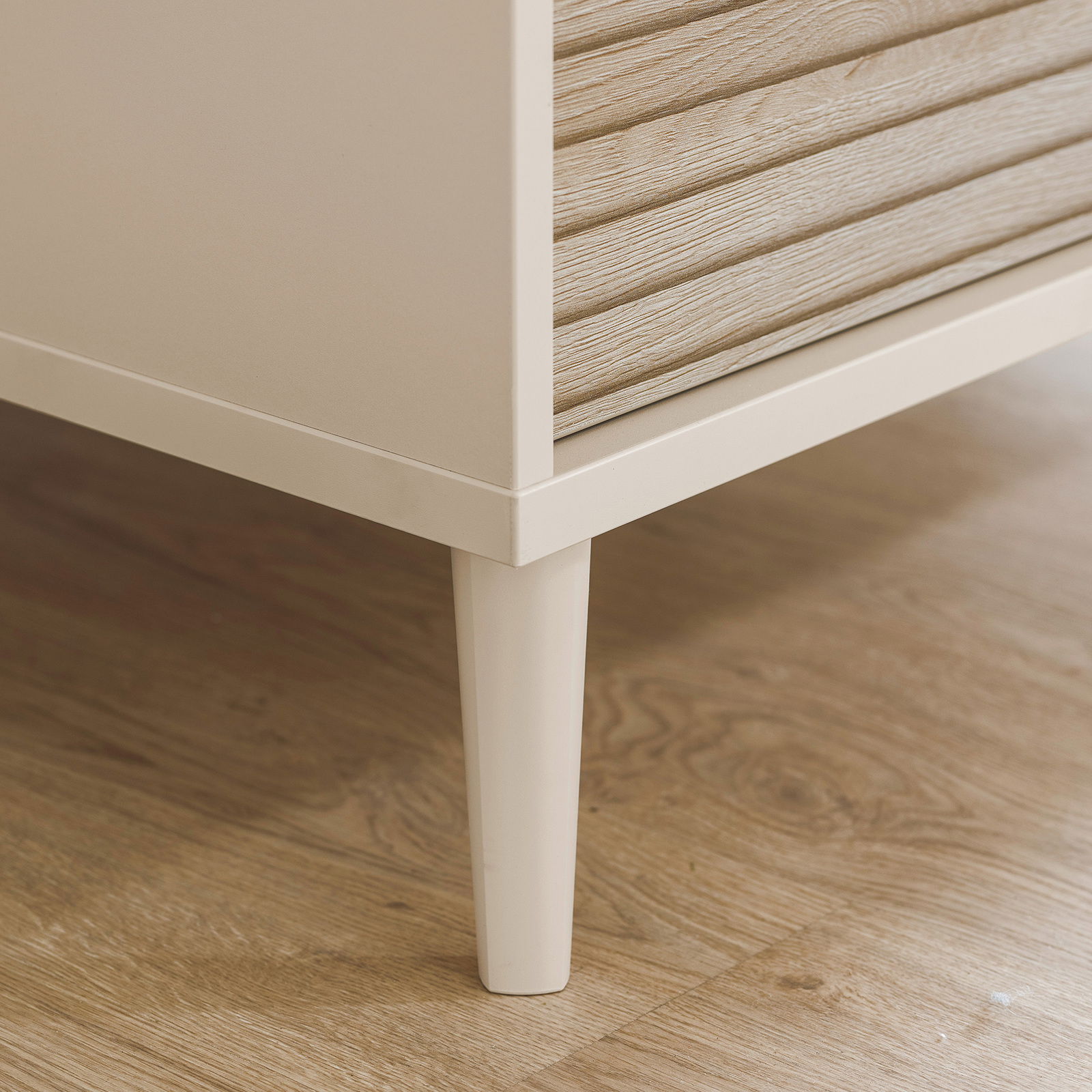 Nahaufnahme der unteren Ecke eines Kleiderschranks mit Holzoptik und weißen Beinen, auf einem Holzboden stehend.