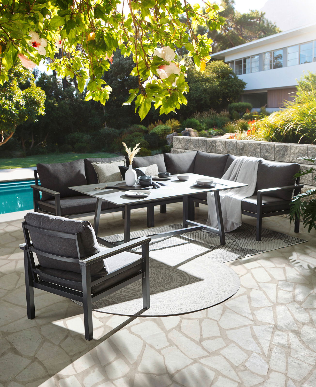 Lounge-Set im Garten mit Blick auf einen Pool, bestehend aus einem Ecksofa, einem Tisch und einem Sessel, umgeben von grüner Natur.