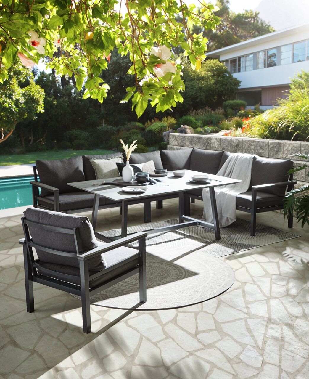 Loungeset in de tuin met uitzicht op een zwembad, bestaande uit een hoekbank, een tafel en een fauteuil, omgeven door weelderig groen.