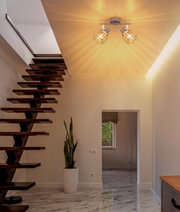 Moderne Deckenleuchte mit zwei verstellbaren Spotköpfen und goldfarbenen Drahtschirm-Käfigen, warm leuchtend im Flur über einer Holztreppe, fotografiert aus leichtem Untersichtwinkel zur Decke
