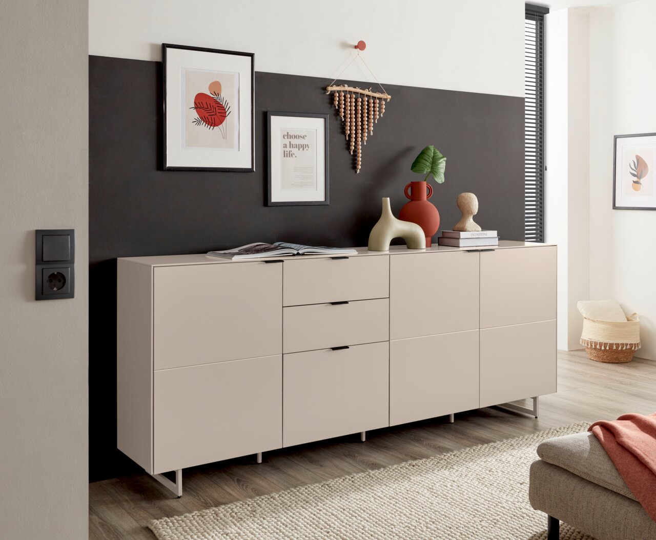 Modern dressoir in greige/beige met drie deuren en twee lades op slanke metalen poten, versierd met vazen en schilderijen, in schuin vooraanzicht in de woonkamer.