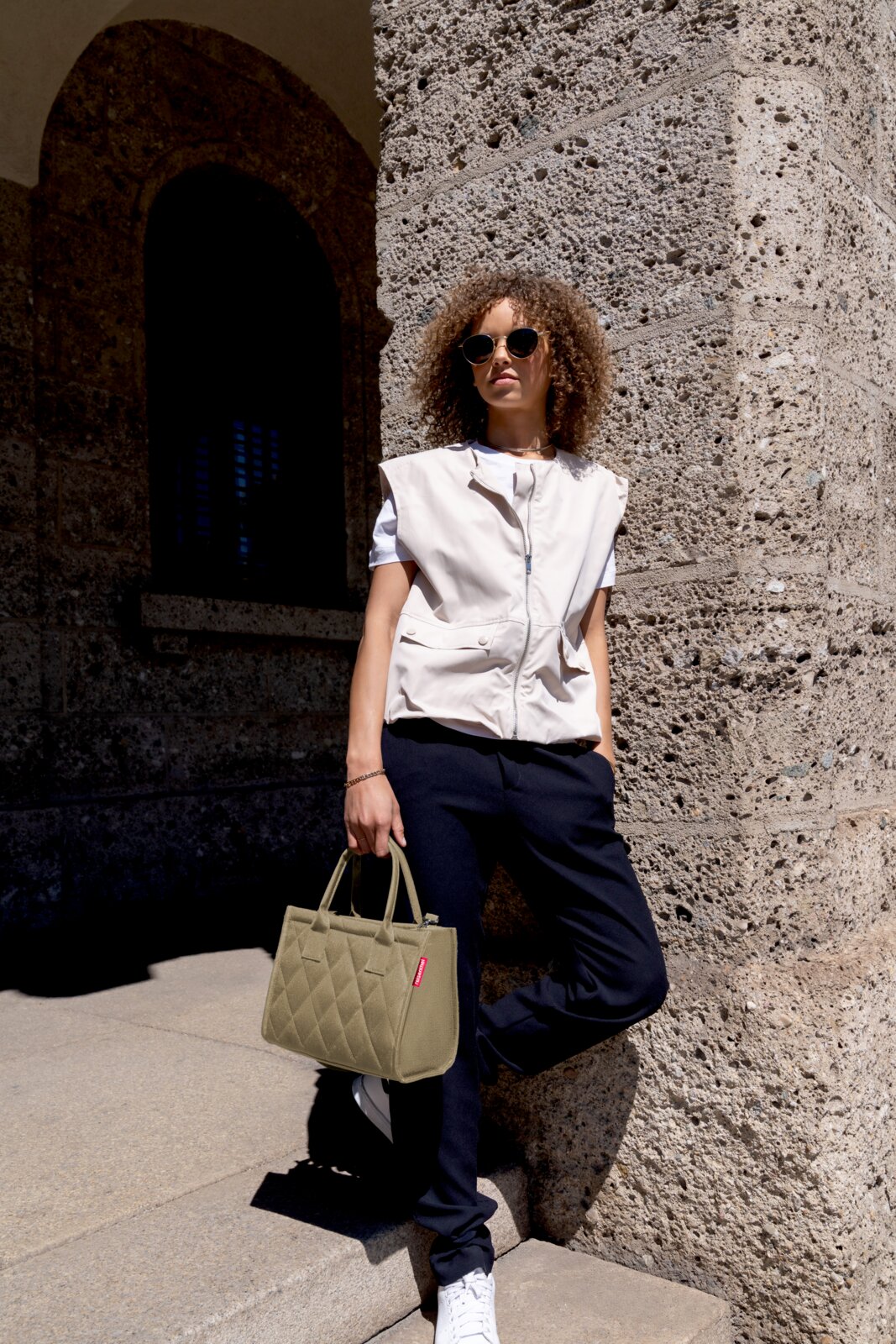 Frau mit Sonnenbrille lehnt an einer Steinwand und hält eine olivgrüne, gesteppte Handtasche in der Hand. Die Tasche ist im Vordergrund sichtbar.