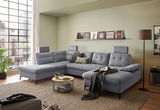 Graues Ecksofa DURBAN in U-Form mit verstellbaren Kopfstützen, dekoriert mit Kissen, in einem modernen Wohnzimmer mit großen Fenstern und grüner Wand, aus der Perspektive von vorne links.