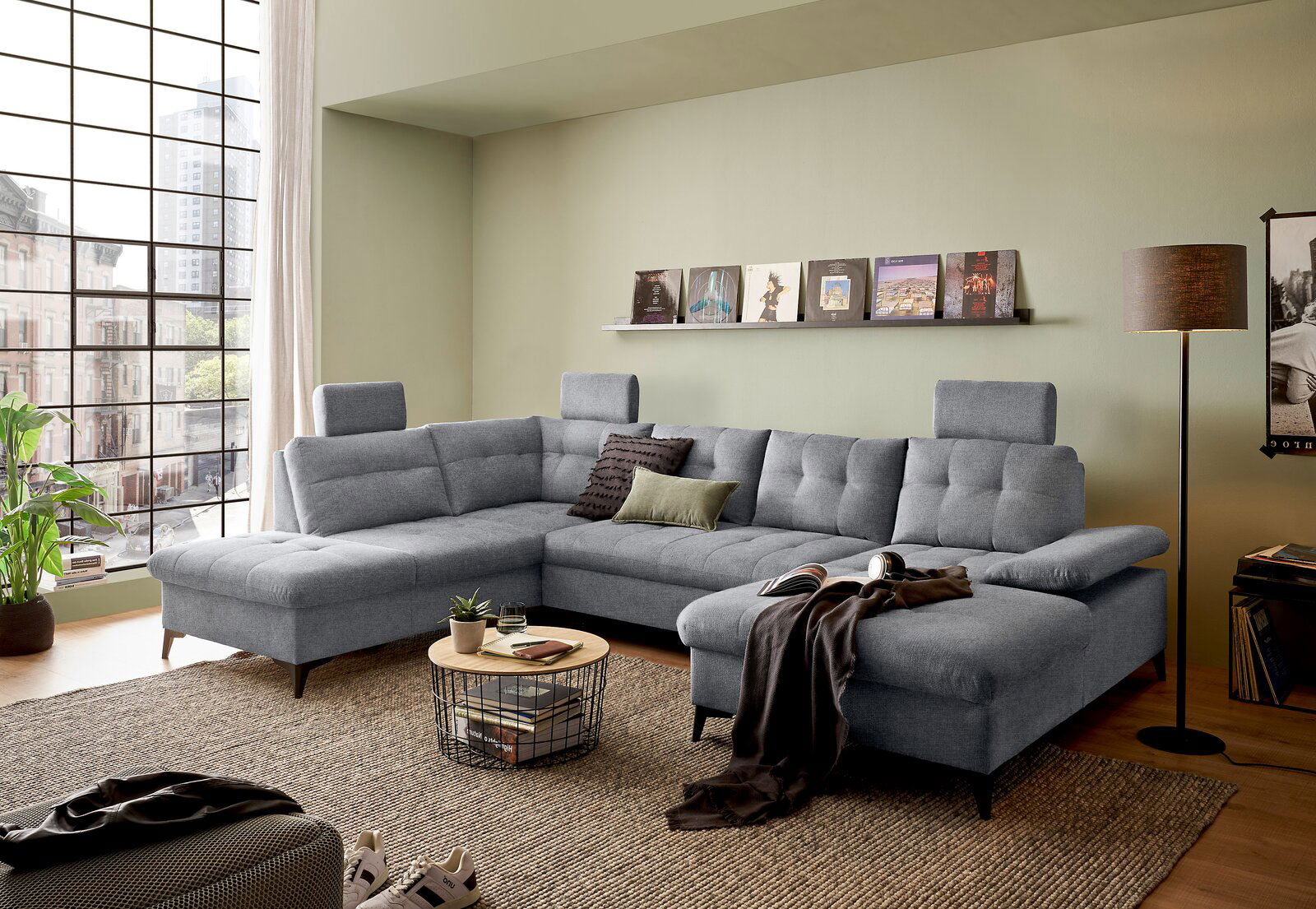 Graues Ecksofa DURBAN in U-Form mit verstellbaren Kopfstützen, dekoriert mit Kissen, in einem modernen Wohnzimmer mit großen Fenstern und grüner Wand, aus der Perspektive von vorne links.