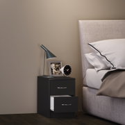 Schwarzer Nachttisch mit zwei Schubladen (untere geöffnet) neben einem gepolsterten grauen Bett, darauf eine moderne Tischlampe, ein Bilderrahmen und ein kleiner runder Wecker; Perspektive: seitliche Frontansicht auf Augenhöhe.
