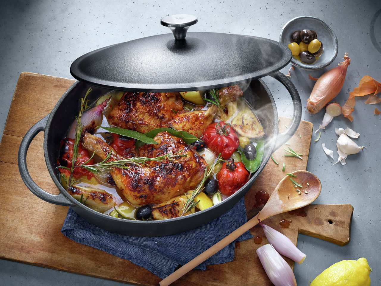 Gusseiserner Bräter mit Deckel, gefüllt mit gebratenem Hähnchen, Tomaten, Zwiebeln und Kräutern, aus der Vogelperspektive fotografiert.